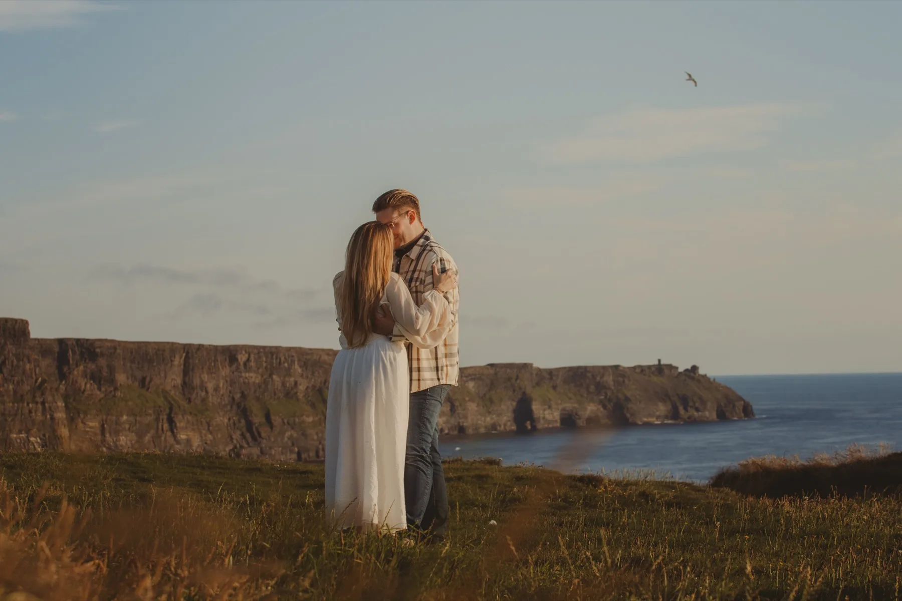 Shannon and Oscar kissing in a grassy field above the sea.