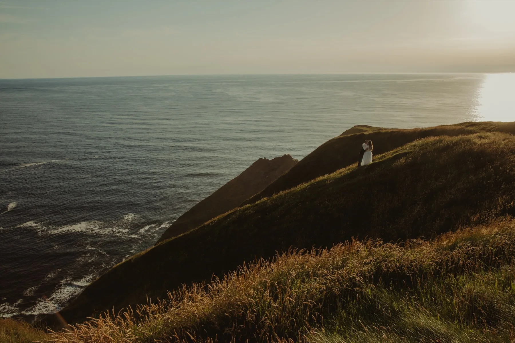 Shannon and Oscar standing together on a grassy cliff above the sea at golden hour.