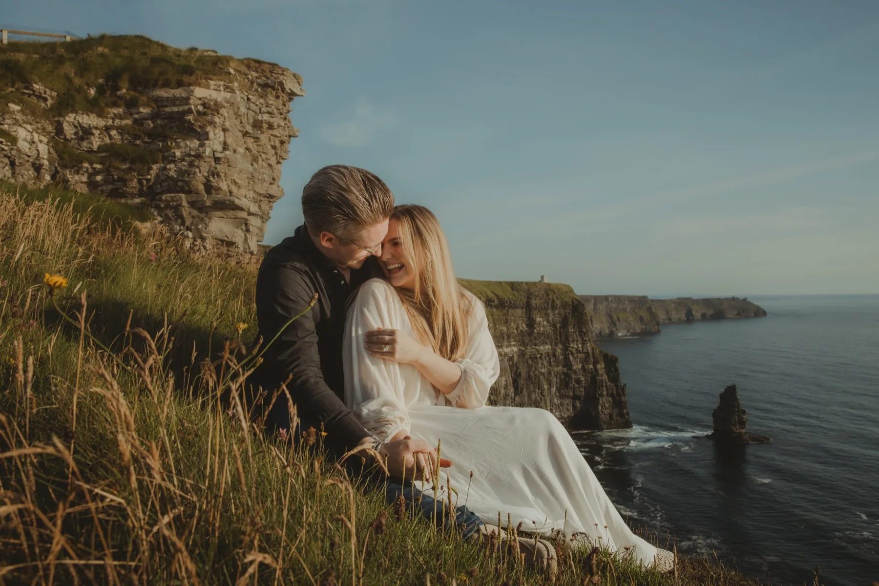 Shannon and Oscar sitting close together in the grass on the edge of a cliff, laughing, with the sea behind them.