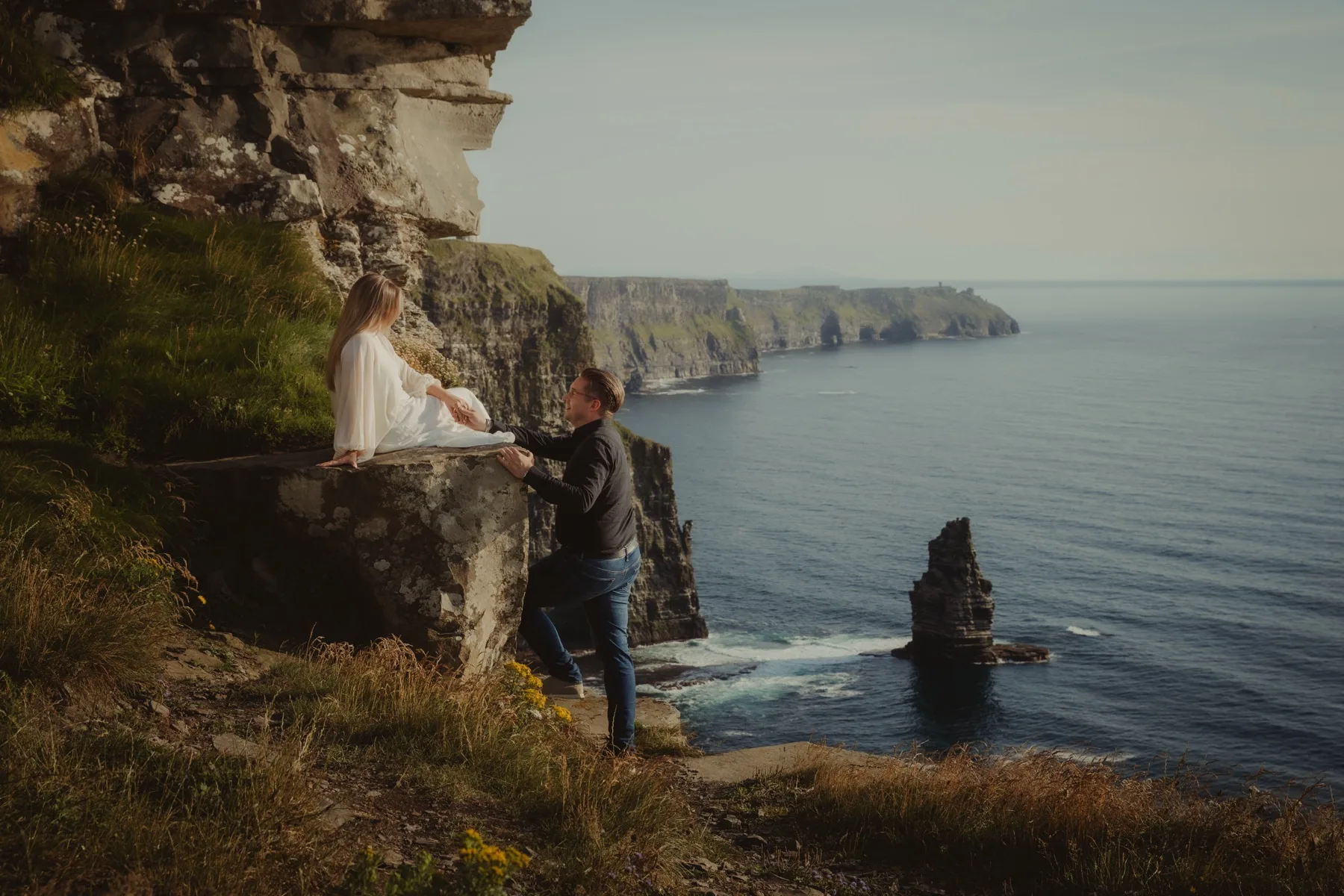 Shannon sitting on a cliff ledge with Oscar standing below her, the Irish coastline stretching out behind them