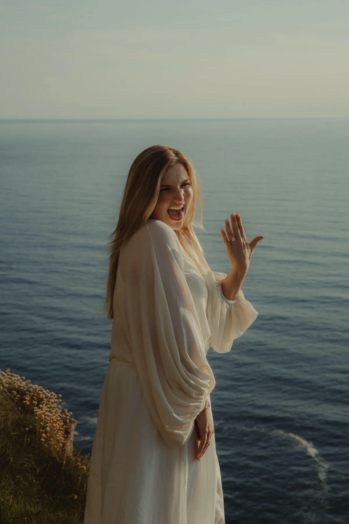 Shannon laughing and waving, showing her engagement ring, with the sea behind her.